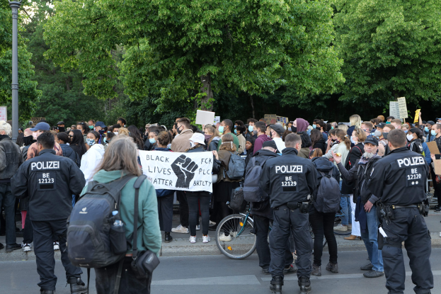 Eine große Gruppe von Menschen steht an der Straße, einige halten Schilder, mit einem Fahrrad im Vordergrund und Bäumen und einem Mast im Hintergrund, an einer Black-Lives-Matter-Demonstration in Berlin teilnehmend.