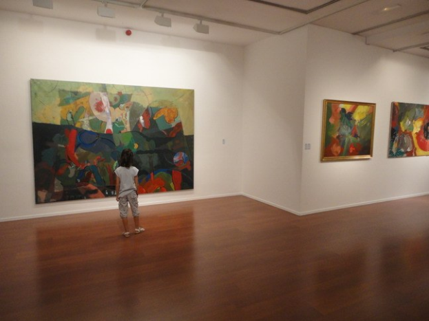 Ein junges Mödchen in einer Kunstgalerie betrachtet gerahmte Bilder an den Wänden, umgeben von stehenden Besuchern unter warmen Deckenlichtern.