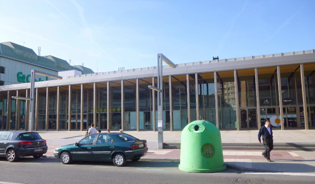 Grüner Müllcontainer an der Seite einer Straße neben einem Glaswandgebäude mit Säulen, mit Fahrzeugen und Fußgängern in der Nähe, unter einem bewölkten Himmel am Berliner Hauptbahnhof.