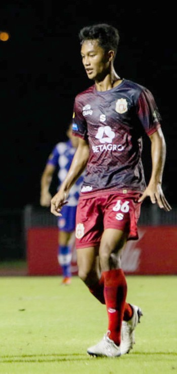 Ein Mann in einem rostroten Hemd und roten Shorts spielt Fußball auf einem Feld, mit einer anderen Person im Hintergrund und einer Tafel mit Schrift.
