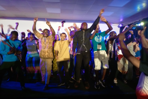 Eine Gruppe von Menschen, die auf einer Bühne tanzen und ihre Hände in die Luft strecken, beleuchtet von Scheinwerfern auf der rechten Seite und einem Bildschirm im Hintergrund, unter einer Decke mit Scheinwerfern, die eine Partyvorstellung suggerieren.