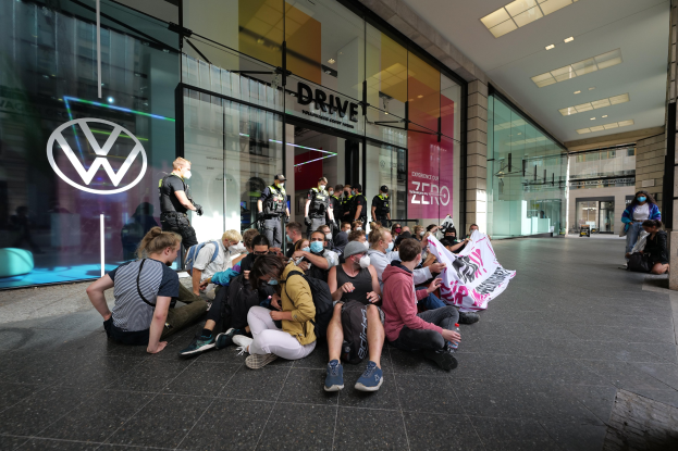 Eine Gruppe von Menschen sitzt vor einer Volkswagen-Verkaufsstelle mit Glaswänden auf dem Boden und hält ein Transparent mit Text, unter Deckenleuchten, scheinbar protestierend.
