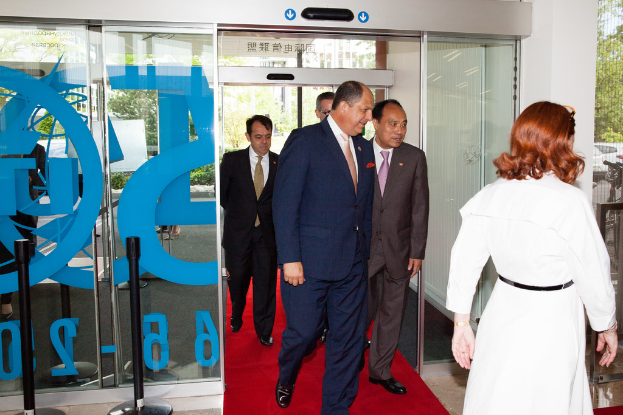 Eine Frau in einem weißen Kleid steht rechts auf einem roten Teppich, umgeben von ein paar Männern; links sind Sicherheitsstangen und eine Glaswand zu sehen, mit Bäumen im Hintergrund; im Hintergrund rechts fahren Fahrzeuge auf der Straße, mit Bäumen und Himmel darüber.