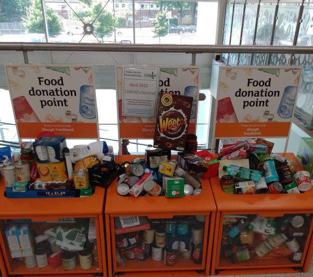 Orange Schübe mit Lebensmittelspenden, darunter Flaschen, Dosen und Kisten, mit informativen Schildern im Hintergrund, vor einem Fenster mit Blick auf Fahrzeuge, Bäume, Gebäude und Himmel.