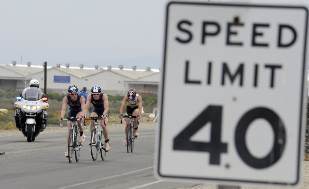 Gruppe von Radfahrern mit Helmen, die eine Straße mit einem Geschwindigkeitsbegrenzungsschild entlangfahren, Gebäude und Bäume im Hintergrund unter einem klaren blauen Himmel.