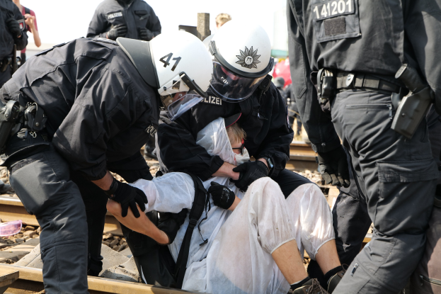 Eine Gruppe von Polizisten in Einsatzausrüstung verhaftet einen Mann in einem weißem Kleid auf einem Bahngeleis, mit anderen im Hintergrund.