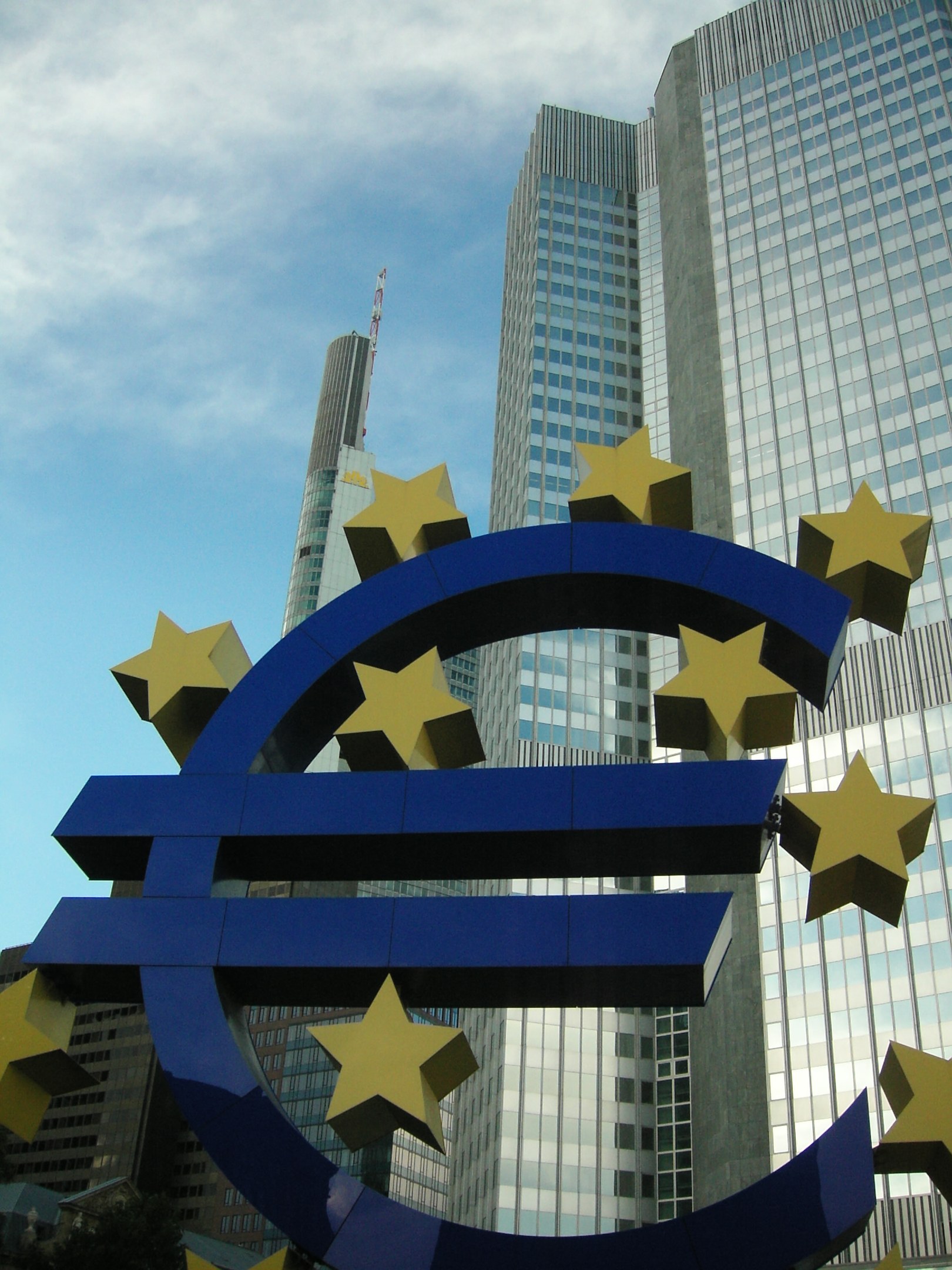 Eurozeichen vor dem Europäischen Zentralbank-Hauptsitz in Frankfurt, Deutschland, mit dem Gebäude umgeben von Bäumen unter einem bewölkten Himmel.