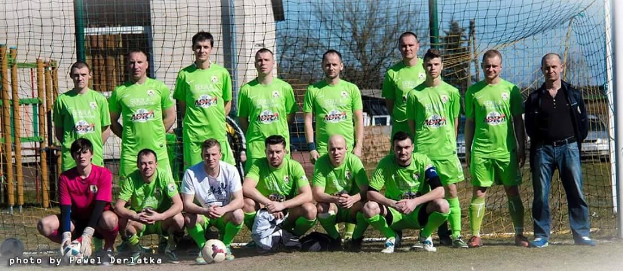 Eine Gruppe von Männern in grünen T-Shirts steht auf einem Fußballfeld, einige sind in der Hocke, mit einem Ball und einem Torpfosten im Hintergrund.