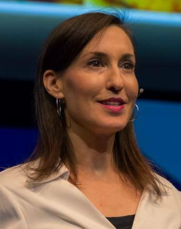 Frau in weißer Jacke und schwarzem Oberteil, das vor einem blauen und gelben Hintergrund sitzt und Ohrringe und ein Mikrofon trägt.