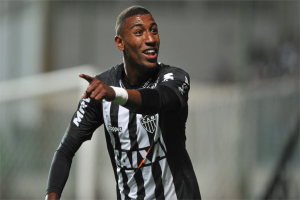 Ein Fußballspieler in einem schwarzen und weißen T-Shirt lächelt und feiert nach einem Tor, mit einem unscharfen Hintergrund.