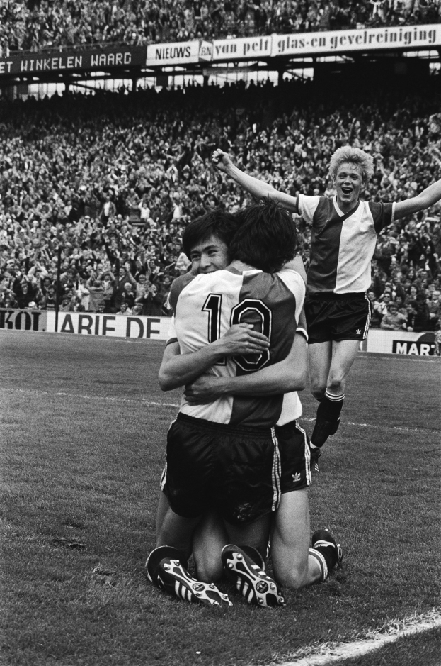 Drei Füßballspieler umarmen sich auf dem Boden nach einem Tor, mit jubelnden Fans und schwenkenden Fahnen in einem schwarz-weßen Stadion Setting.