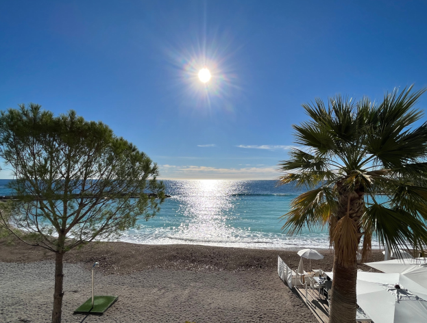 Ein Strand mit Palmen, Sonnenschirmen und üppiger Vegetation unter einem strahlend blauen Himmel mit der Sonne im Hintergrund.