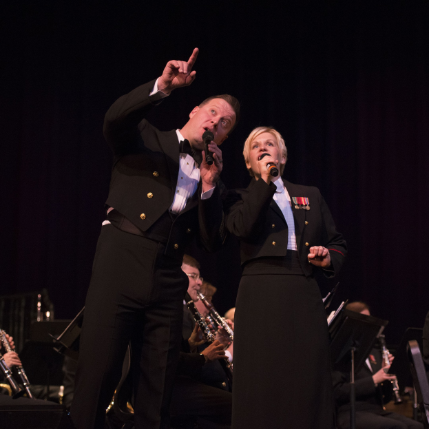 Ein Mann und eine Frau in Fracks mit Mikrofonen auf einer Bühne, mit einem sitzenden Orchester dahinter in einer schwach beleuchteten Umgebung.