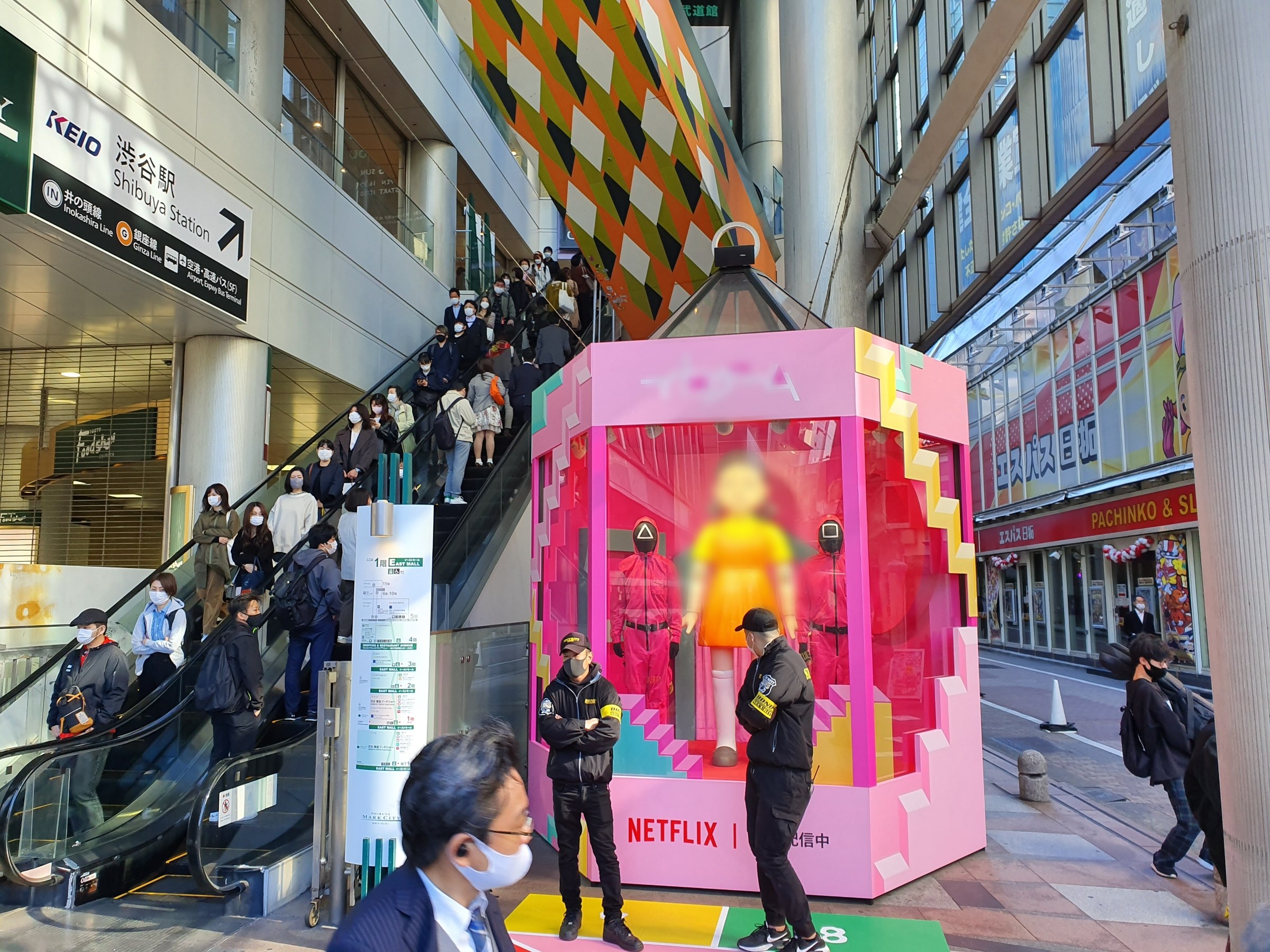 Eine Gruppe von Menschen steht vor einem Gebäude mit Glaswänden und Texttafeln, einige tragen Masken, während eine Rolltreppe mit Menschen zu sehen ist. In der Mitte steht ein pinker Kasten mit der Aufschrift "Netflix's new Netflix-Booth in Tokyo".