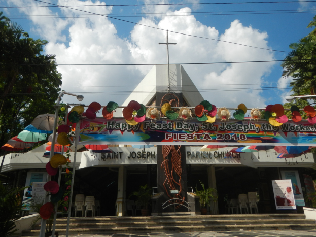 Ein Gebäude mit der Aufschrift "Frohes Fest St. Josef der Arbeiter" über dem Eingang, mit Treppen, einem dekorierten Rundbogen, Außenbestuhlung mit Tischen und Stühlen, Topfpflanzen, flankierenden Bäumen und einem bewölkten Himmel.