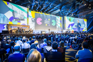 Eine große Menschenmenge sitzt auf Stühlen vor einer Bühne bei der Deutschen Internetkonferenz 2019, mit einem Tisch voller Gegenstände links und Bildschirmen und Lichtern im Hintergrund.