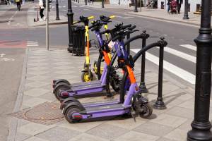 Eine Reihe von Elektro-Scootern, die entlang einer Straße mit Fußgängern, Radfahrern, einem Müllcontainer im Vordergrund und Gebäuden, Bäumen und anderen Objekten im Hintergrund geparkt sind.