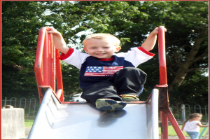 Ein Kind rutscht eine Rutsche hinunter, mit einem Zaun am Ende, einem anderen Kind rechts unten und Bäumen im Hintergrund.