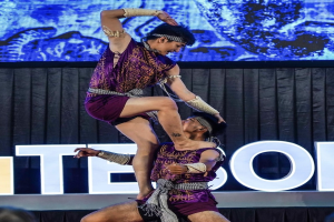 Zwei Performer in purpurfarbenen Kleidern führen einen akrobatischen Hebemanöver auf der Bühne vor einem Publikum aus, mit einem schwarzen Vorhang und einem Bildschirm im Hintergrund und sichtbaren Zuschauern unten.