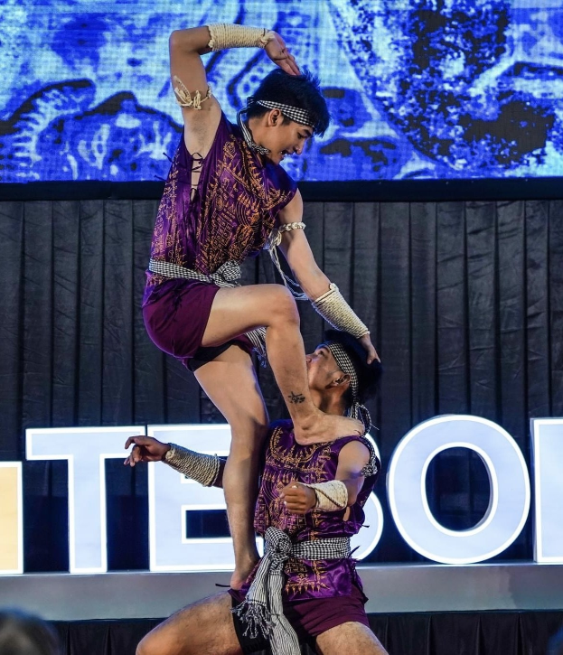 Zwei Performer in purpurfarbenen Kleidern führen einen akrobatischen Hebemanöver auf der Bühne vor einem Publikum aus, mit einem schwarzen Vorhang und einem Bildschirm im Hintergrund und sichtbaren Zuschauern unten.