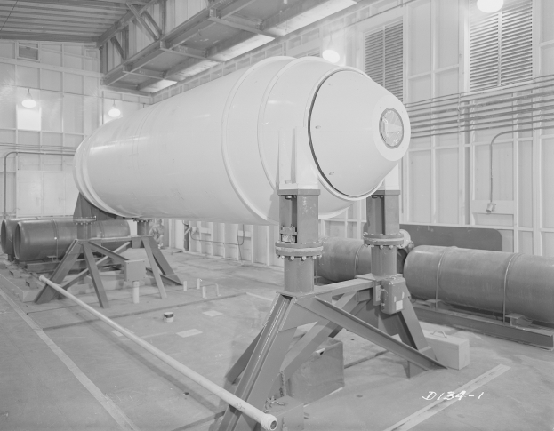 Große zylindrische Wasserstoffbombe auf einem Ständer in einer industriellen Fabrikumgebung mit Rohren, Lampen und einer Wand im Hintergrund.