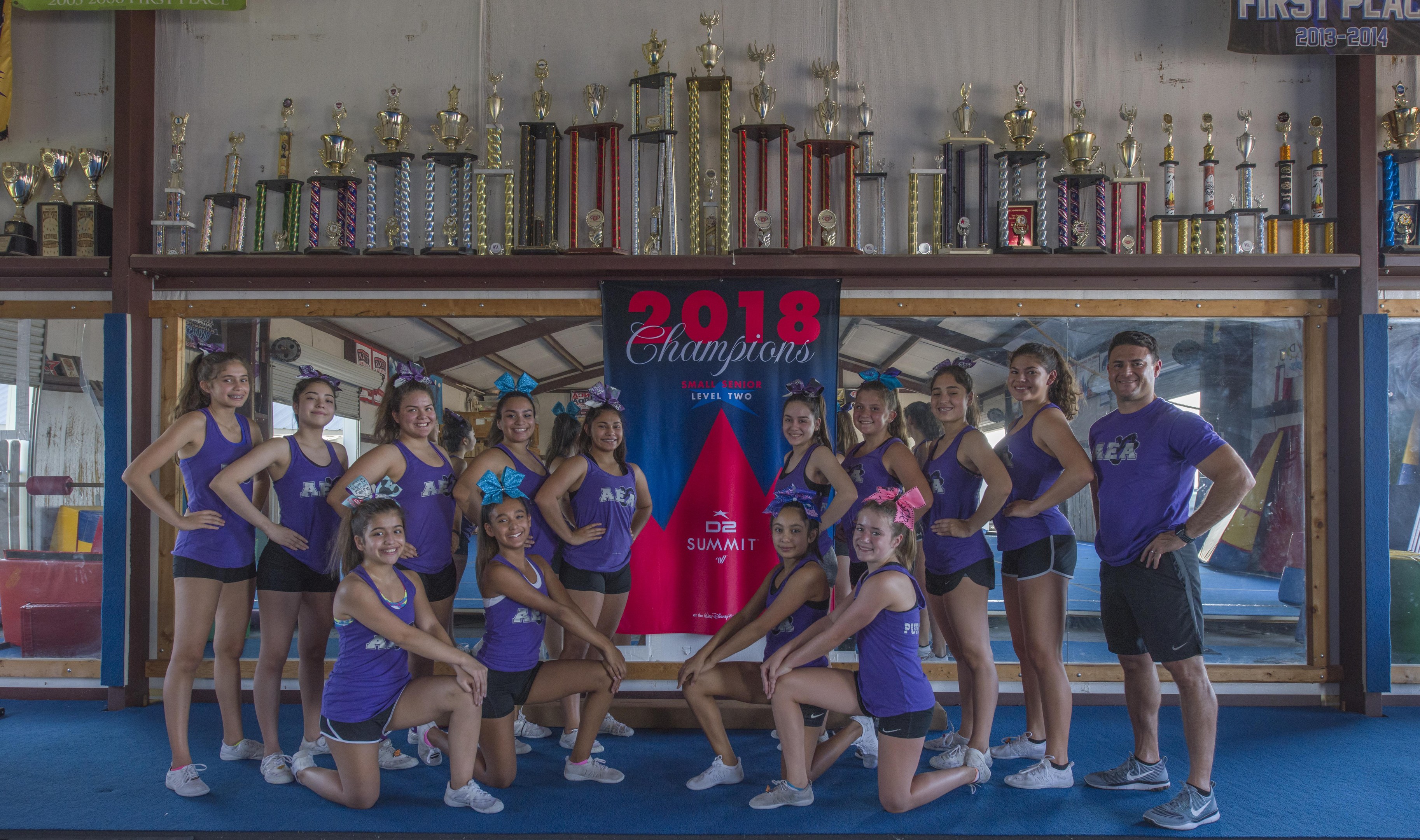Gruppe von Cheerleadern in lila T-Shirts und schwarzen Shorts, die vor einemvitrine mit Trophäen posieren, im Hintergrund ein Banner mit der Aufschrift "2018 Summit Cheerleading Champions".
