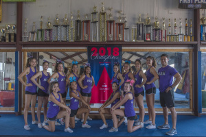 Gruppe von Cheerleadern in lila T-Shirts und schwarzen Shorts, die vor einemvitrine mit Trophäen posieren, im Hintergrund ein Banner mit der Aufschrift "2018 Summit Cheerleading Champions".