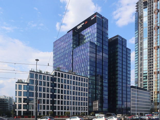 Großes modernes Gebäude mit zahlreichen Fenstern, identifiziert als Deutsche Bank Hauptsitz in Frankfurt, Deutschland, umgeben von städtischer Infrastruktur einschließlich Straßenlaternen, Fahrzeugen, Bäumen und einem bewölkten Himmel.