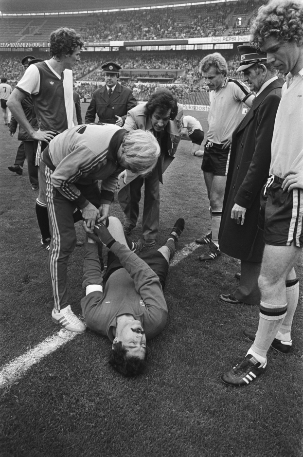 Schwarzes und weißes Foto eines Fußballspielers, der auf dem Boden liegt und von einer Gruppe Männer und einer Stadionmenge umgeben ist, wobei er verletzt zu sein scheint.