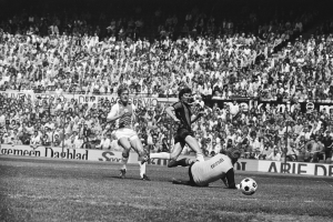 Schwarzes Foto von Fussballspielern auf einem Feld während eines Bundesliga-Spiels zwischen Ajax und Hannover 96 in der Saison 1978-79, mit Zuschauern im Hintergrund.