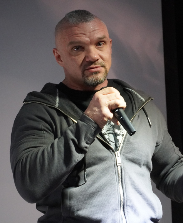 Ein Mann in einer schwarzen Kapuzenjacke und einem schwarzen Anzug mit einem weißen Hemd, der ein Mikrofon hält, steht selbstbewusst vor einer Wand und sieht aus wie ein Komiker.