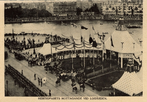 Ein altes Schwarz-Weiß-Foto einer Menge, die sich um einen Karneval mit Zelten, Fahnen, Stangen und Pferden im Vordergrund und Gebäuden, Bäumen und Wasser im Hintergrund versammelt hat, mit Text am unteren Bildrand.
