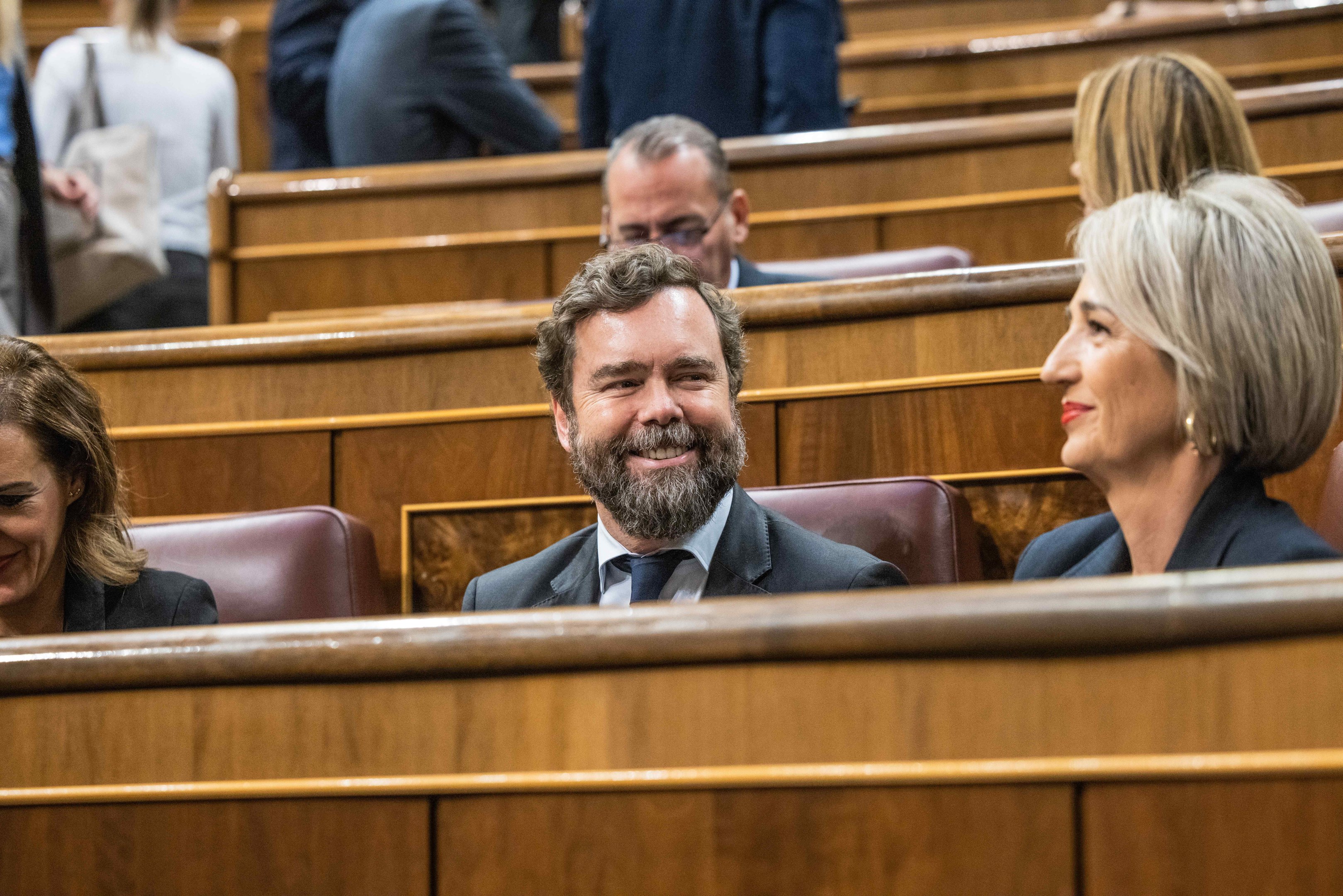 Eine Gruppe von Menschen, die in einem Gerichtssaal sitzen, mit einem Mann in Anzug und Krawatte und zwei Frauen in legerer Kleidung, die nebeneinander sitzen, sowie Bänke im Hintergrund.