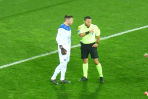 Ein Schiedsrichter in einem gelben Hemd und schwarzen Shorts spricht mit einem Fussballer in einer weissen und blauen Uniform auf dem Feld, beide tragen Turnschuhe.