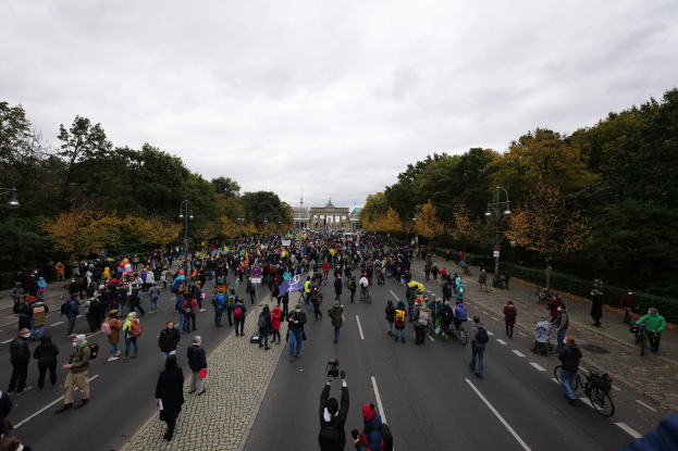 Eine große Gruppe von Menschen marschiert auf einer von Bäumen gesäumten Straße mit Laternenmasten, einem Gebäude im Hintergrund und einem klaren Himmel, einige halten Kameras während einer Demonstration in Berlin.