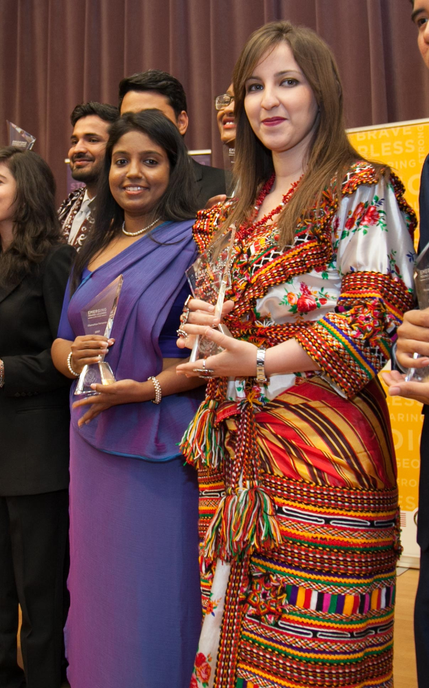 Eine Gruppe von Menschen auf einer Bühne mit einer Frau in der Mitte, die eine Trophäe hält, hinter einem Vorhang mit einer Tafel, auf der 'Bravo Blessing Award Gewinner' steht.