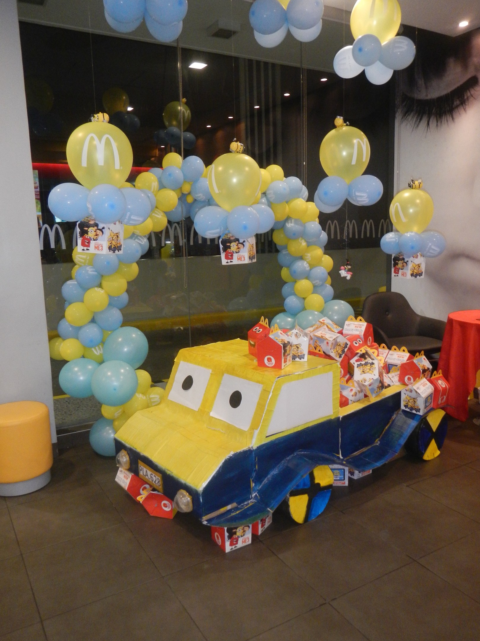 Eine McDonald's-Geburtstagsfeier mit einem gelben und blauen Lastwagen, der mit Ballons und anderen Dekorationen geschmückt ist, einem roten Tisch mit Gegenständen, Hockern, einer Glaswand und Deckenleuchten, die eine festliche Atmosphäre schaffen.