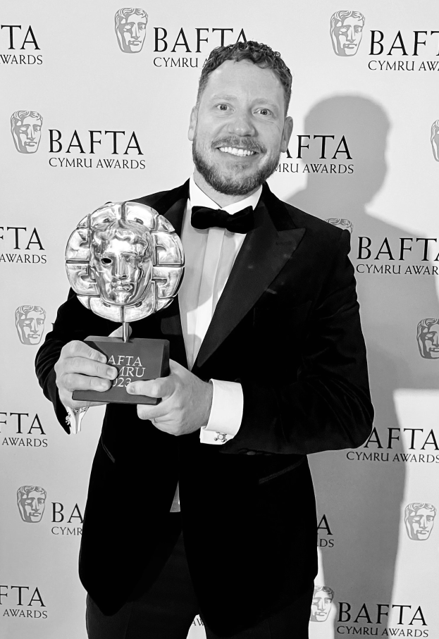 Ein Mann in einem Smoking, der eine BAFTA-Trophäe hält und lächelt, mit einem Hintergrundschild, auf dem "BAFTA Awards 2018" steht.