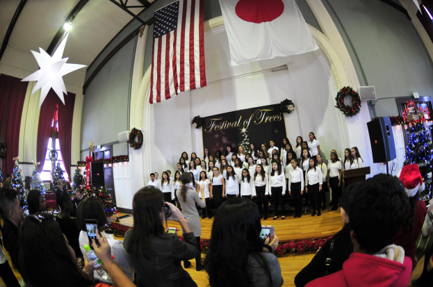 Eine Gruppe von Menschen auf einer Bühne mit einem Mikrofon, Lautsprechern, geschmückten Weihnachtsbäumen, einer Tafel mit Text und einer Menge mit Kameras und Handys.