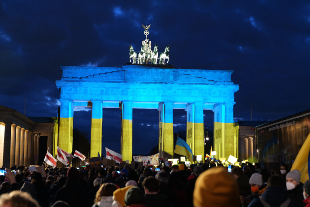 Menge mit Flaggen und Schildern vor dem Brandenburger Tor in Berlin, mit einem Banner auf der rechten Seite mit protestrelevanten Text.