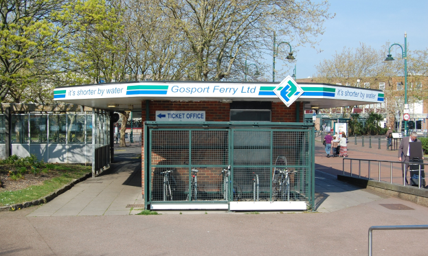 Haltestelle mit einem 'Gosport Ferry Ltd.'-Schild, Metallzaun, Menschen, Fahrrädern, Bäumen, Straßenlaternen, Schildern, einem Gebäude und einem bewölkten Himmel.