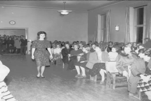Eine Frau in einem schwarzen und weißen Kleid läuft über den Laufsteg während einer Modenschau für die Frauenwahlrecht, mit einer Menge, die auf Stühlen sitzt und einer Uhr und Lampen an der umliegenden Wand.