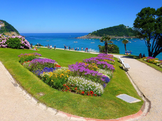 Ein Park mit bunten Blumenbeeten, umgeben von grünem Gras und Bäumen, mit Bänken, Fußwegen, Menschen, Booten auf dem Wasser, Gebäuden und einem klaren blauen Himmel im Hintergrund.