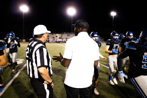 Ein Fußballtrainer in einer Mütze spricht zu seinem Team auf einem Feld bei Nacht, mit Spielern in Helmen, Laternenmasten und einem dunklen Himmel im Hintergrund.