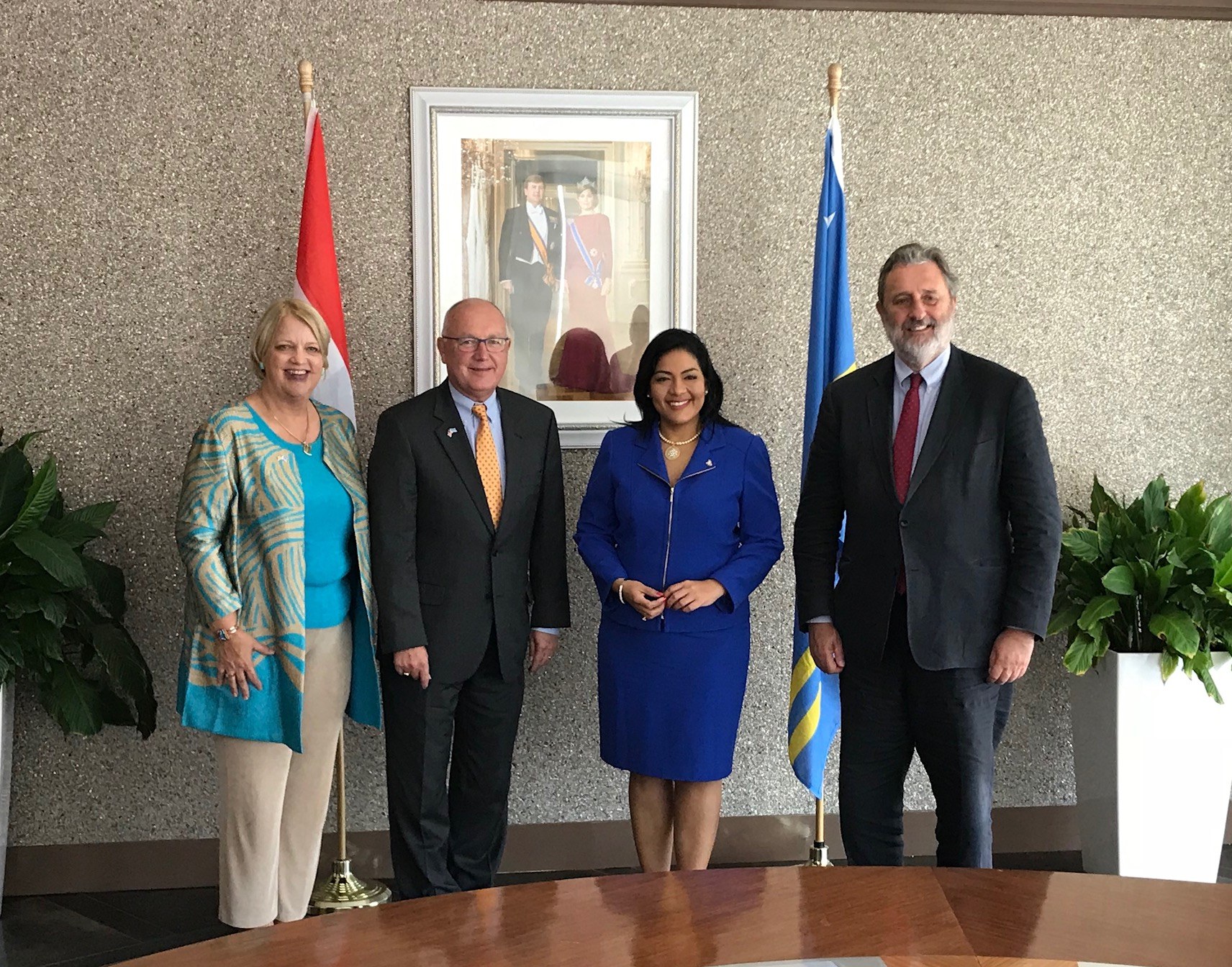 Eine Gruppe von Menschen, die um einen Tisch mit Papieren stehen und von Flaggen, Topfpflanzen und einem Rahmenfoto an der Wand umgeben sind.