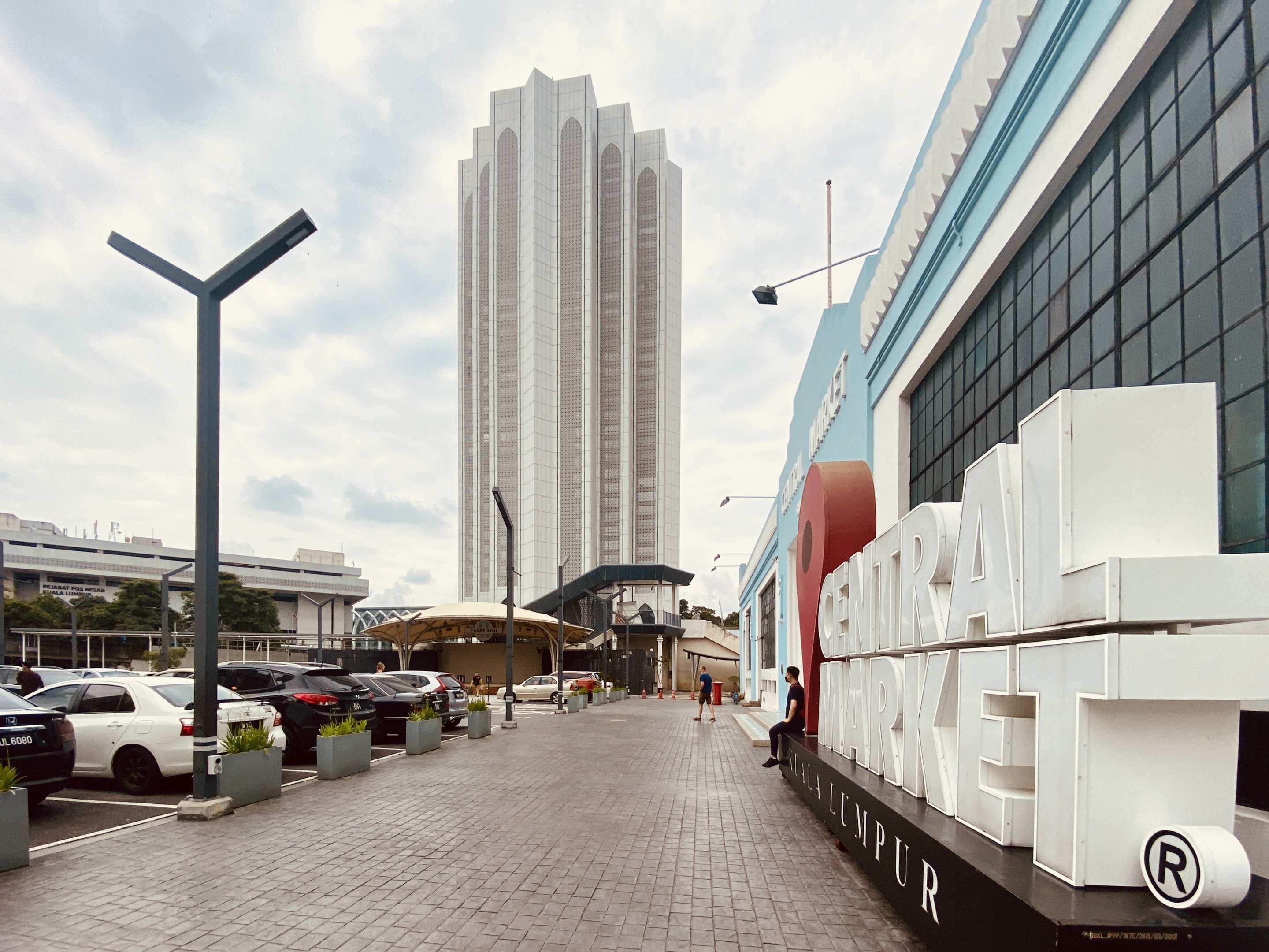 Ein belebter zentraler Markt in Kuala Lumpur, Malaysia, mit hohen Gebäuden, Straßenlaternen, Straßenlaternen, Kraftfahrzeugen, Fußgängern, Namensschildern, Bäumen und einem bewölkten Himmel.