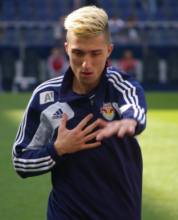Ein junger Mann in einem blauen T-Shirt steht auf einem Fußballfeld und streckt die Arme aus, zeigt eine entschlossene Miene und hat einen unscharfen Hintergrund.