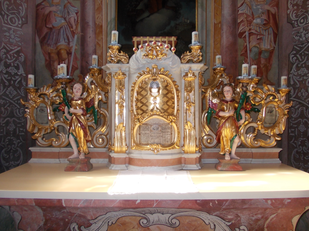 Ein Altar in einer Kirche mit Skulpturen von Menschen und Kerzen auf St"nden, vor einer Wand mit einem Gemälde, das eine feierliche Atmosph"are schafft.