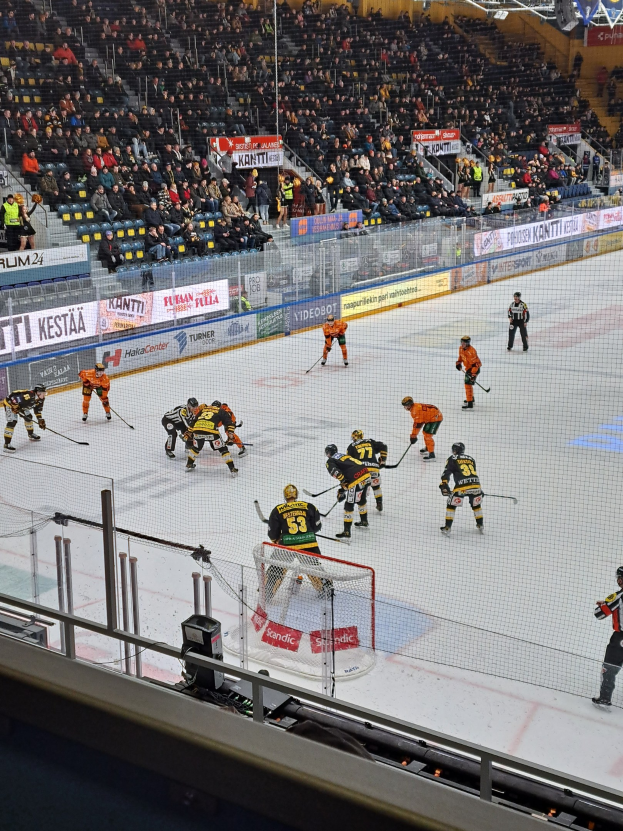 Eine Gruppe von Menschen, die Hockey auf einem Eisring spielt, mit einem Netz und einem Zaun, Zuschauern im Hintergrund und Bannern mit Text, unter Stadionbeleuchtung.