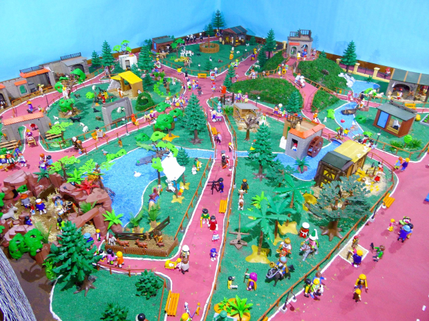 Zoo-Modell mit Miniatur-Leuten, Bäumen, Häusern, Pflanzen, Gras, einem Zaun und einer Hintergrundwand, mit Text unten.